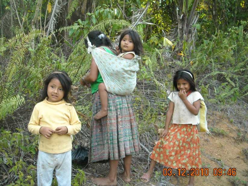 A mother carries a young child in a wrap, with two other young children standing near