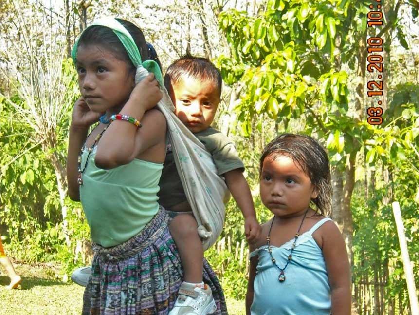A young girl carries a baby in a wrap, while a younger girl stands nearby